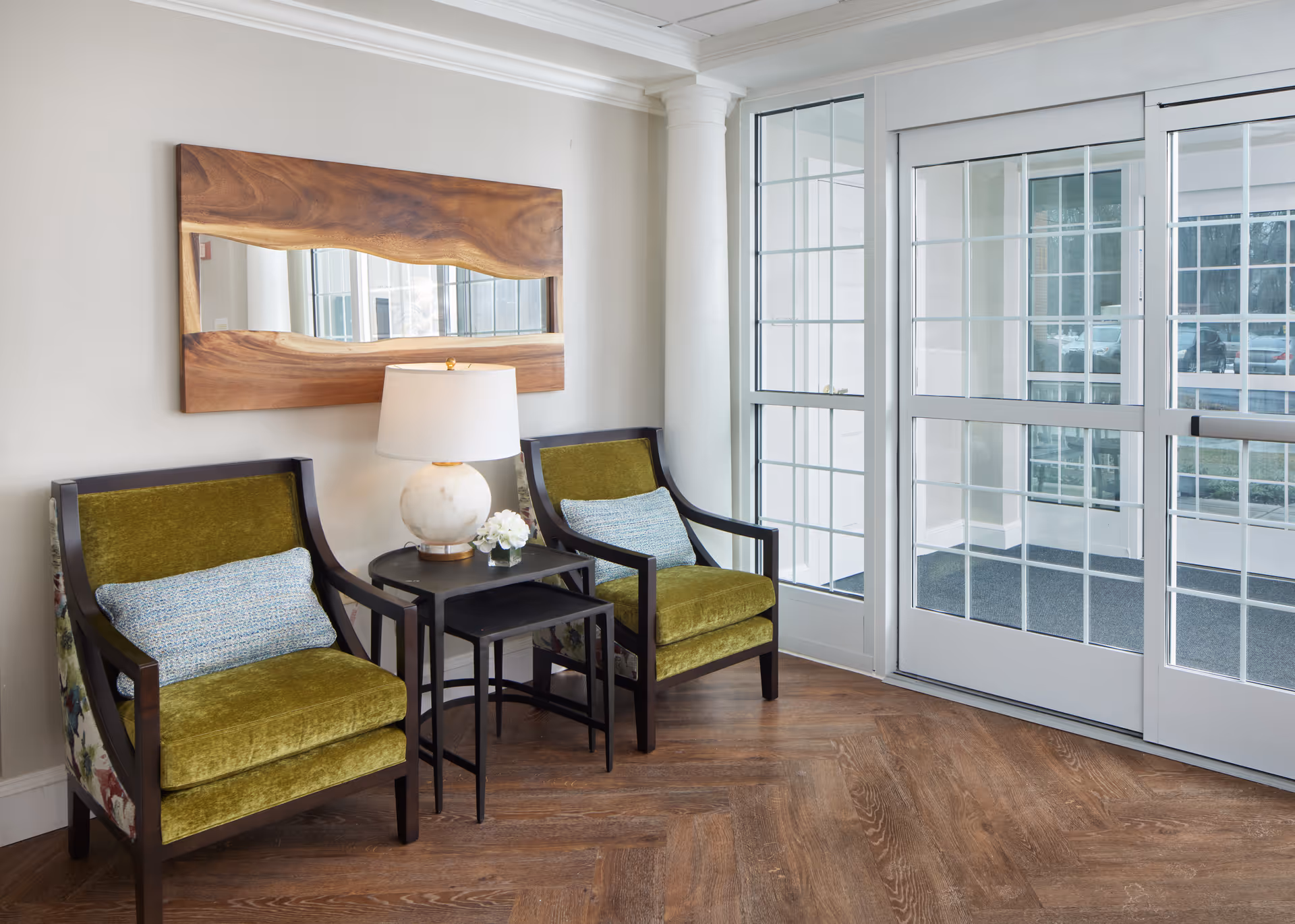 A cozy seating area with two green upholstered armchairs featuring floral patterned sides, separated by two nested black side tables. On the tables are a white marble lamp and a small white flower arrangement. Behind the chairs is a large wooden-framed mirror with a natural edge design. To the right, there are glass double doors with white frames leading outside, letting in natural light. The floor is a warm wood with a herringbone pattern.