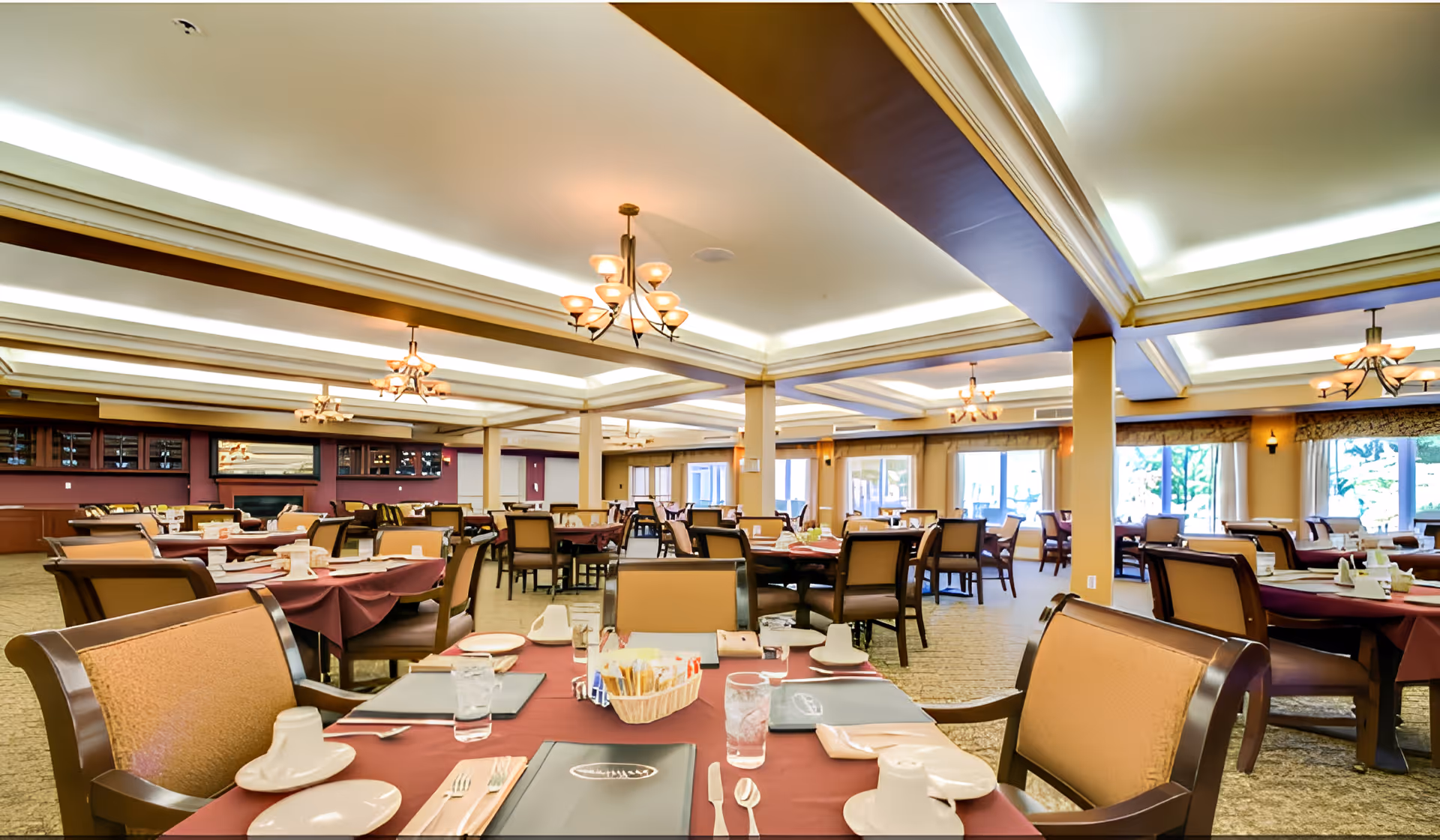 Spacious dining room with multiple tables covered in burgundy tablecloths, set with plates, cups, utensils, and menus. The room features beige cushioned chairs, large windows letting in natural light, and elegant ceiling lights.