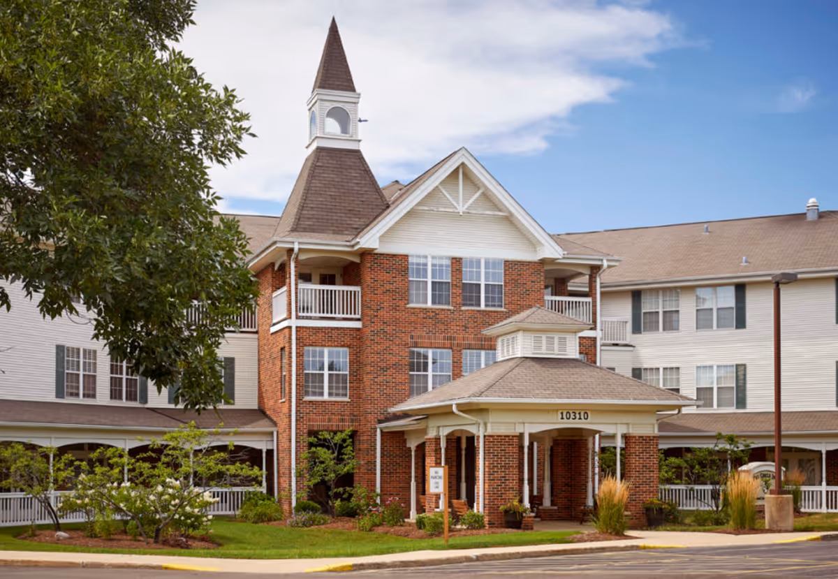 Exterior view of a senior living facility named Peace Village, featuring a multi-story brick and siding building with a peaked roof and a small tower. The entrance has a covered porch with the number 10310 displayed above it. There are trees, shrubs, and a well-maintained lawn in front of the building under a partly cloudy sky.