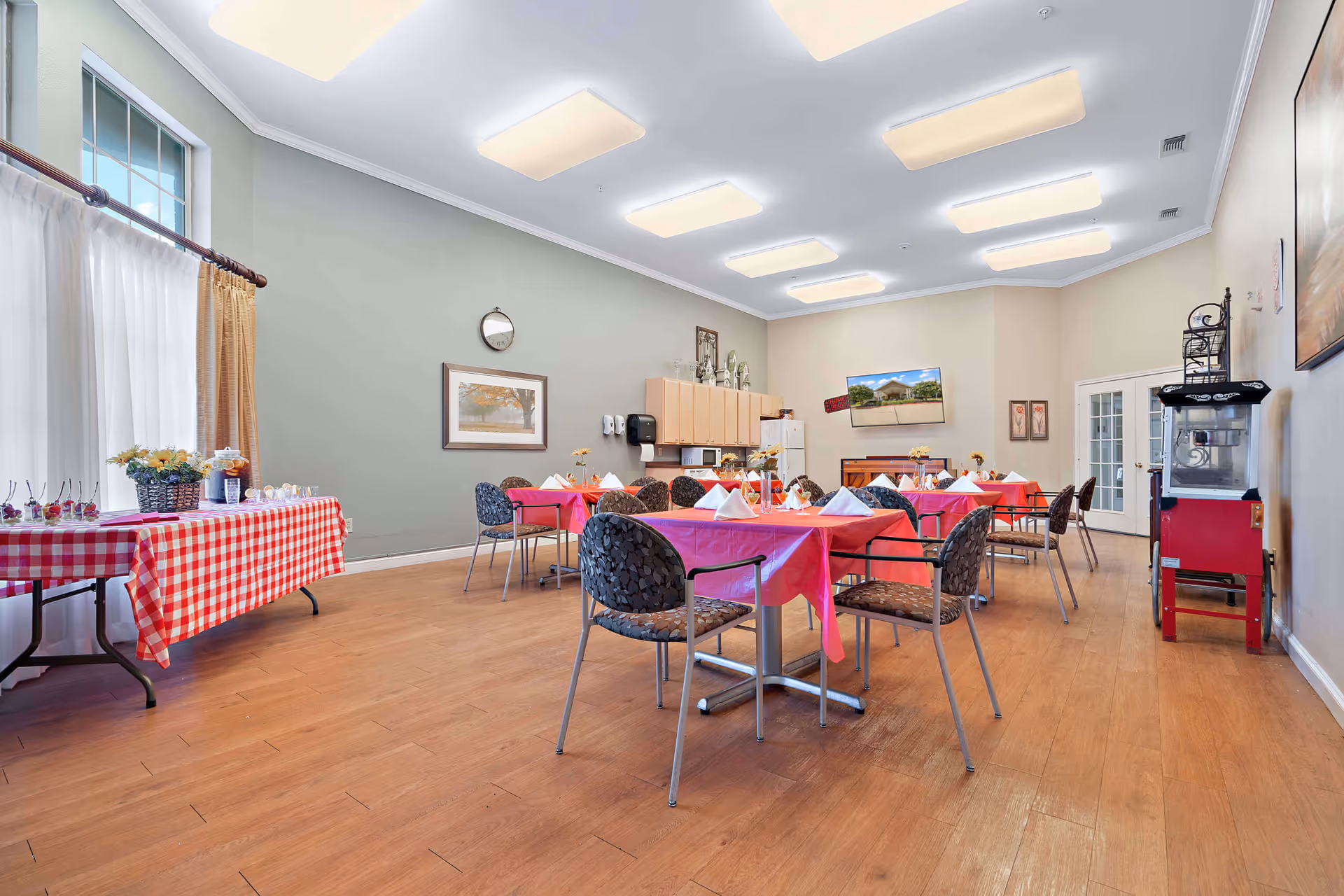 A bright dining room with several tables covered in red and pink tablecloths, each set with white napkins and small flower centerpieces. There are patterned chairs around the tables, a side table with a red and white checkered tablecloth holding snacks and drinks near a window with white curtains. The room has wooden flooring, soft overhead lighting, a wall clock, framed artwork, a television mounted on the wall, and a popcorn machine in the corner.