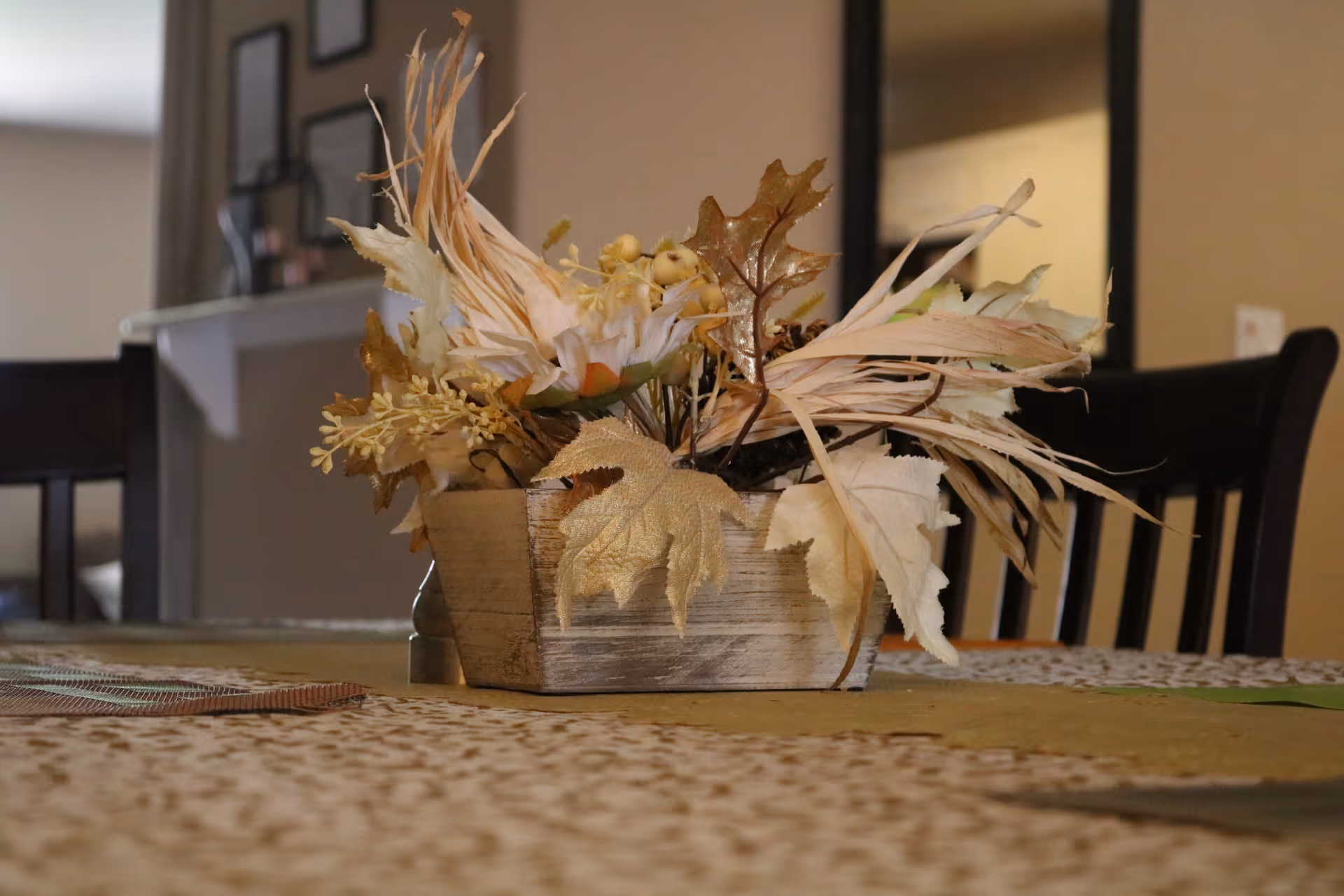 A close-up view of a wooden rectangular planter filled with dried decorative leaves and flowers placed on a table with a patterned tablecloth. In the background, there are dark wooden chairs and a blurred interior setting.