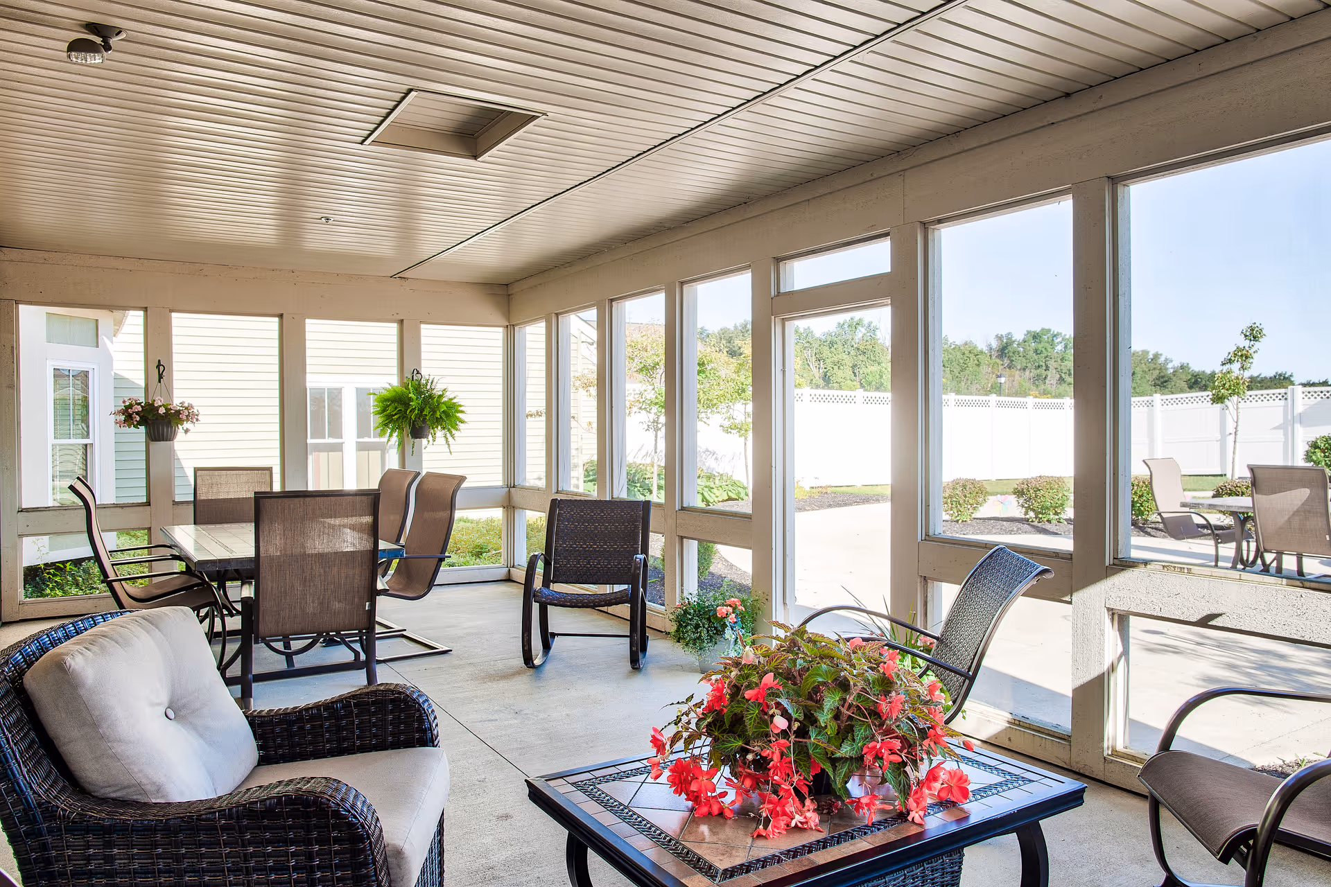 A bright and spacious enclosed patio area with large windows allowing natural light to fill the space. The area is furnished with cushioned wicker chairs, a table with a floral centerpiece, and a dining table with several chairs. Outside the windows, there is a paved patio with additional seating and a white fence enclosing the garden area with shrubs and small trees.