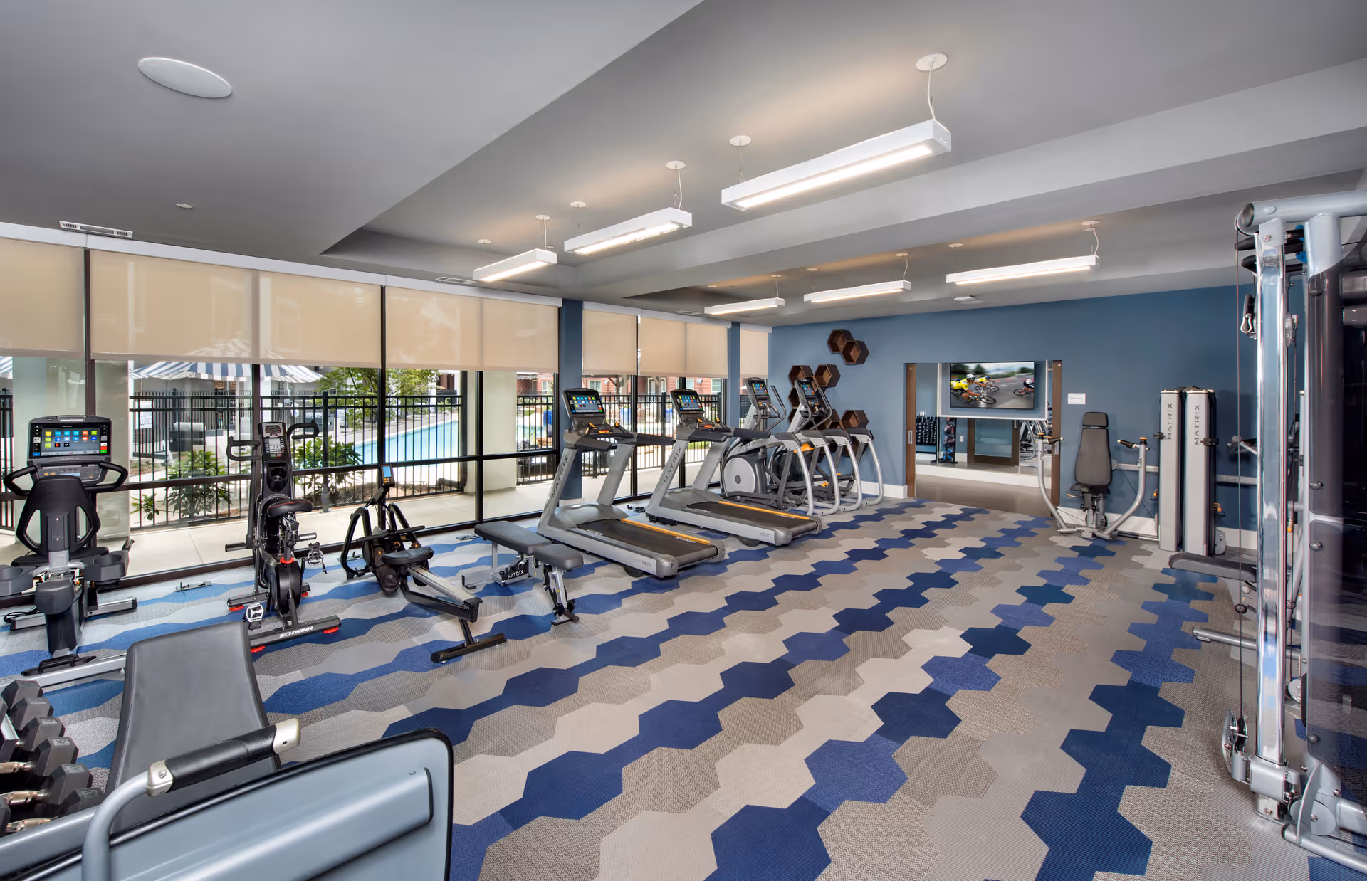 A modern fitness room with various exercise equipment including treadmills, stationary bikes, elliptical machines, weight machines, and benches. The room has large windows with beige roller shades, a patterned carpet in shades of blue and gray, and a blue accent wall with hexagonal wall shelves. A TV is mounted on the far wall above a mirrored section.