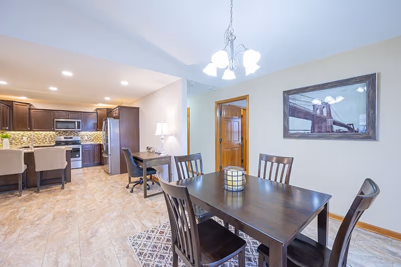 A bright and spacious dining area with a dark wooden table and six matching chairs. A decorative candle centerpiece is on the table. The room features beige tiled flooring and light-colored walls with a framed picture of a bridge. In the background, there is a modern kitchen with dark wood cabinets, stainless steel appliances, and a breakfast bar with two beige stools. A small desk with a lamp and chair is positioned near a wooden door.