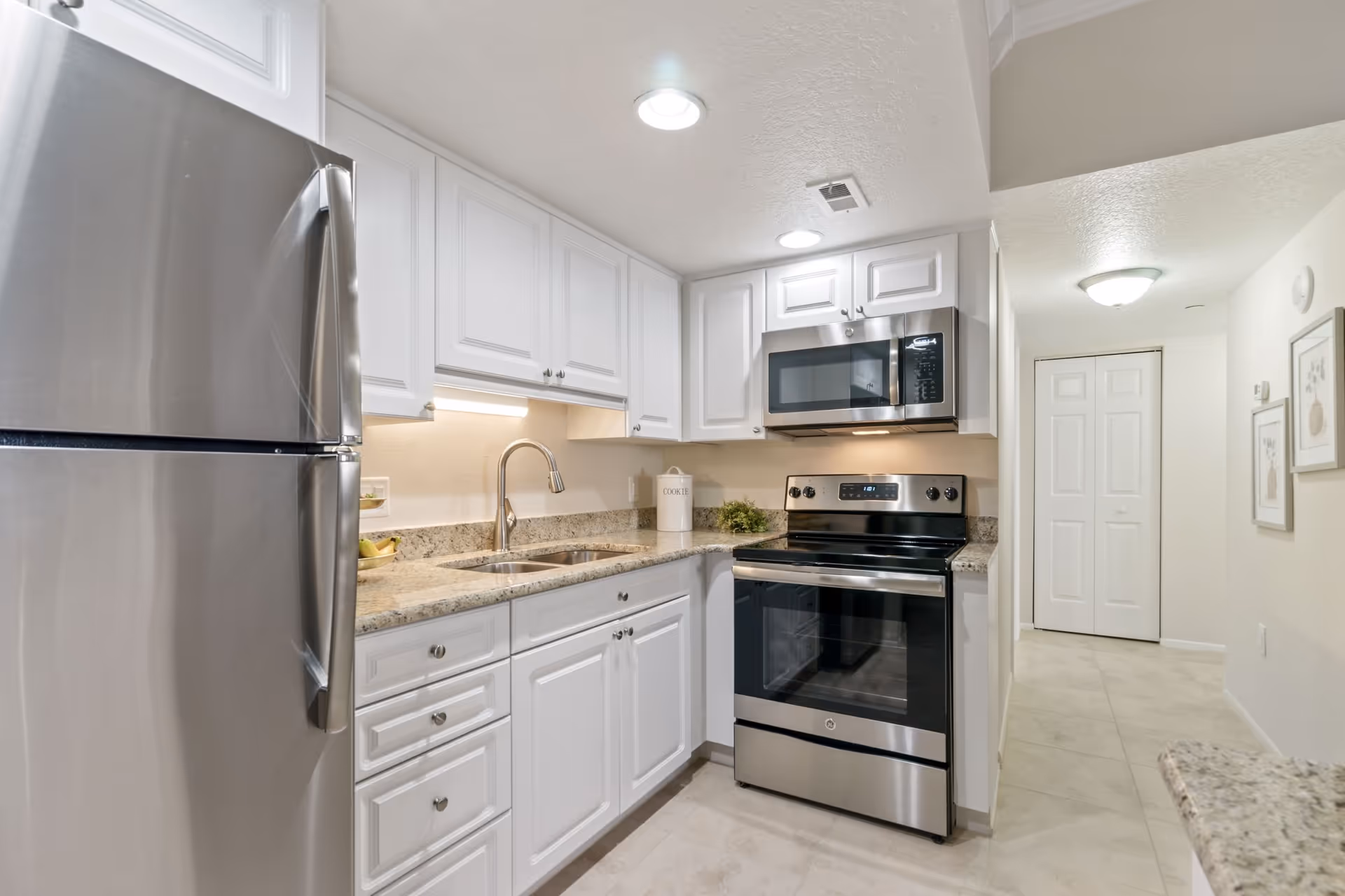 Modern kitchen with white cabinets, granite countertops, stainless steel refrigerator, stove, and microwave. The kitchen is well-lit with recessed ceiling lights and under-cabinet lighting. There is a double sink and a hallway with white doors and framed artwork on the wall.