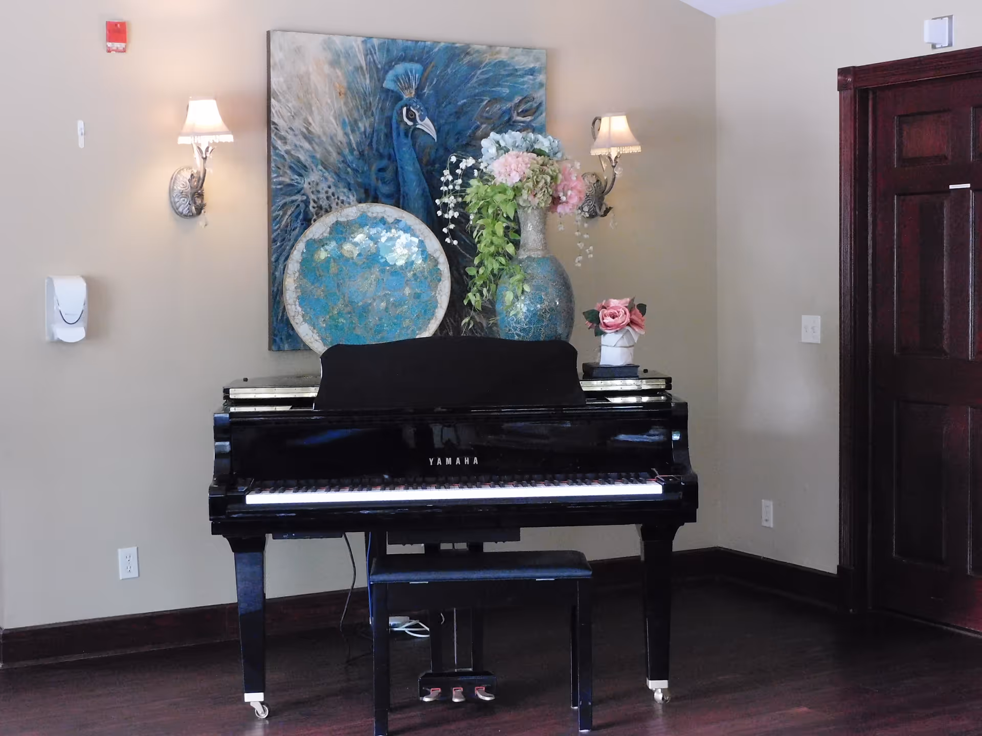 A black Yamaha piano with a matching bench in a room with beige walls and dark wood trim. Above the piano is a large painting of a blue peacock and decorative plates. Two wall-mounted lamps with shades flank the painting. A vase with flowers and a small flower arrangement sit on top of the piano. A dark wooden door is visible on the right side of the image.