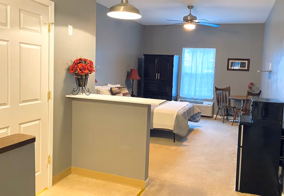 Studio-style living area with a bed, dark armoire, small dining table, and a pass-through counter under pendant lights.