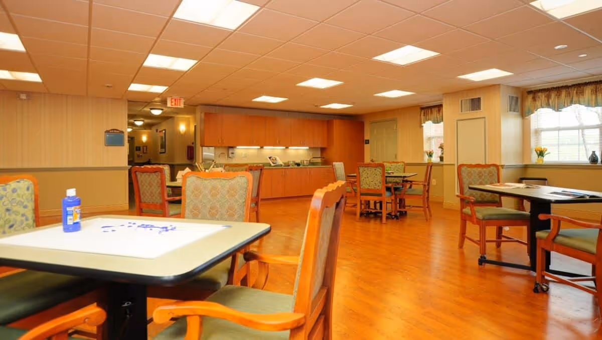 A spacious, well-lit common dining area in a senior living facility with multiple tables and cushioned chairs arranged neatly on a wooden floor. The room features a kitchenette with wooden cabinets and a countertop along the back wall. Large windows with valances allow natural light to enter the room.