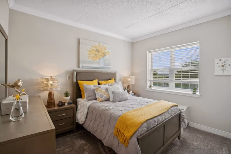 A cozy bedroom with a neatly made bed featuring gray and yellow bedding, two bedside tables with lamps, a dresser with decorative items, a window with blinds letting in natural light, and wall art including a painting of yellow flowers above the bed.