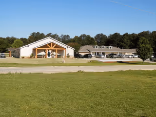 Wide view of Hope Haven Assisted Living Community showing two single-story buildings with pitched roofs, surrounded by a large grassy area and trees in the background under a clear blue sky.