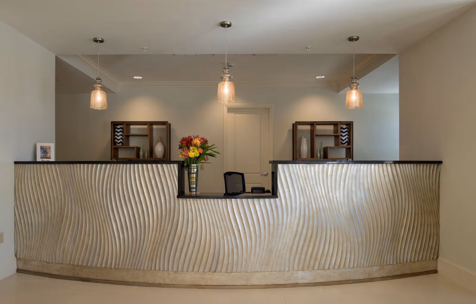 Reception desk area with a textured wave-patterned front panel, three hanging pendant lights above, a vase with colorful flowers on the desk, and two decorative shelving units with vases and art pieces on the wall behind.