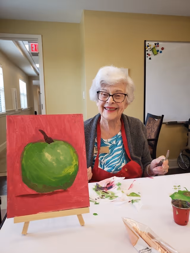 An elderly woman with white hair and glasses is sitting at a table in a room, smiling and holding a painting knife. She is wearing a red apron and a blue patterned shirt. In front of her is an easel displaying a painting of a green apple on a red background. The table has paint stains and a small container of paint. Behind her is a whiteboard with magnets and a chair.