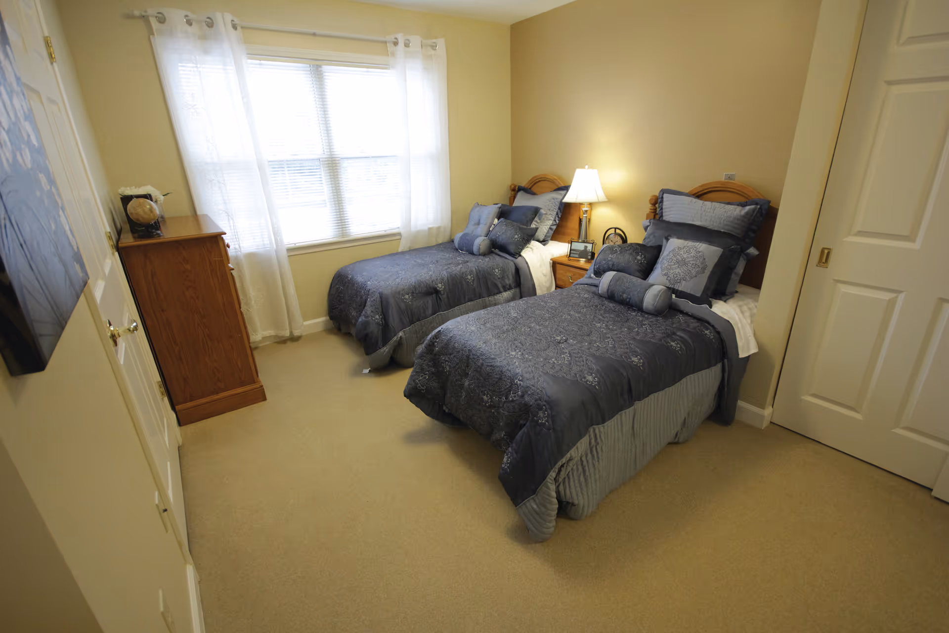 A bedroom with two twin beds dressed in matching blue and gray bedding. Between the beds is a wooden nightstand with a lamp, a clock, and a phone. A window with white curtains lets in natural light. There is a wooden dresser against the wall and a painting hanging on the opposite wall. The room has beige walls and carpeted floor.