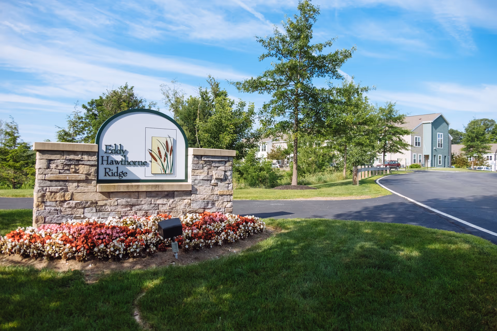 Stone sign for Eddy Hawthorne Ridge surrounded by colorful flowers, with a paved road, green grass, trees, and residential buildings in the background under a blue sky with some clouds.