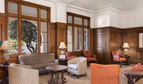 Sunlit senior living lounge with wood-paneled walls, upholstered sofas and chairs, round coffee tables, and lamps by large windows.