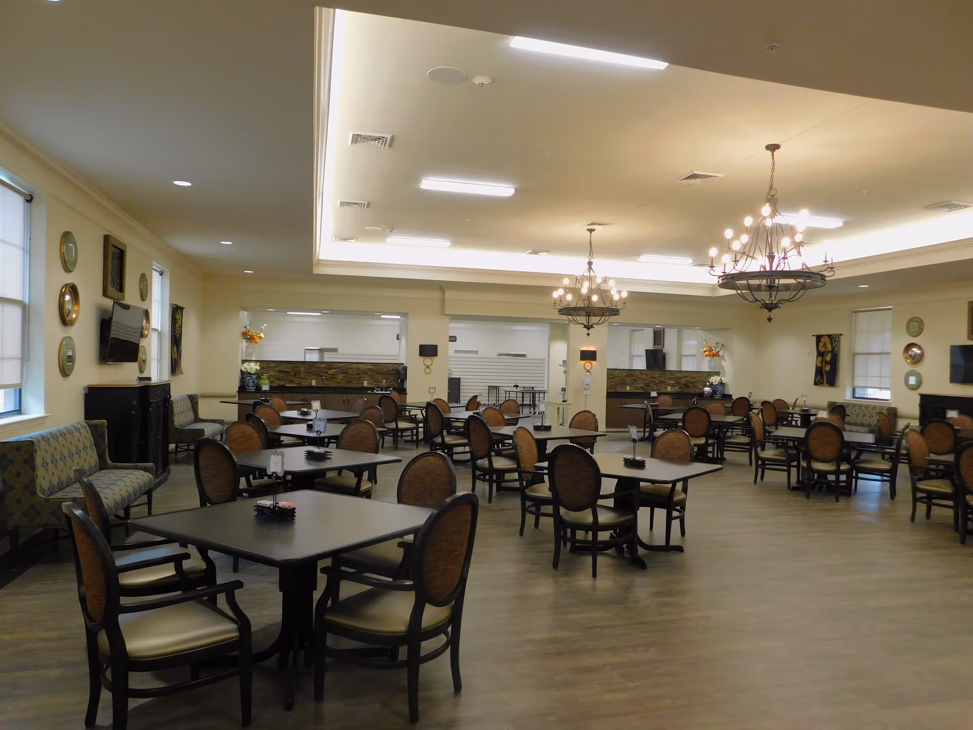 A spacious dining room with multiple square tables and cushioned chairs arranged neatly. The room features two large chandeliers hanging from the ceiling, wall-mounted televisions, decorative plates on the walls, and large windows with blinds. The floor is wooden, and the overall lighting is warm and inviting.