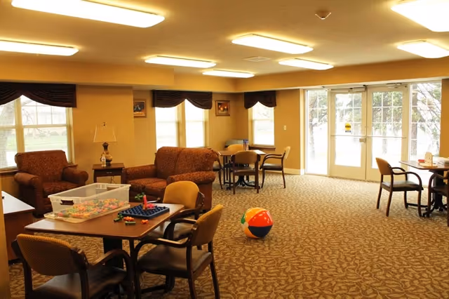 Spacious senior living common room with tables, chairs, sofas, patterned carpet and a colorful beach ball near glass doors.