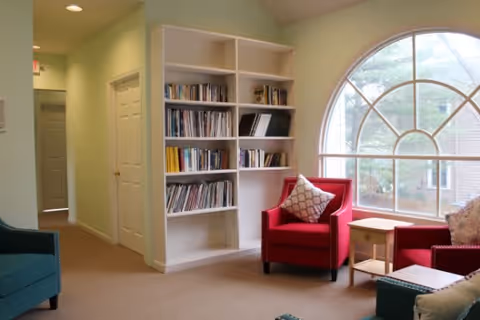 A cozy living room area with a large arched window letting in natural light. The room features a white bookshelf filled with books and magazines, two red armchairs with patterned cushions, a small wooden side table, and part of a teal armchair visible in the foreground. The walls are painted light green and there is a hallway with doors leading to other rooms.