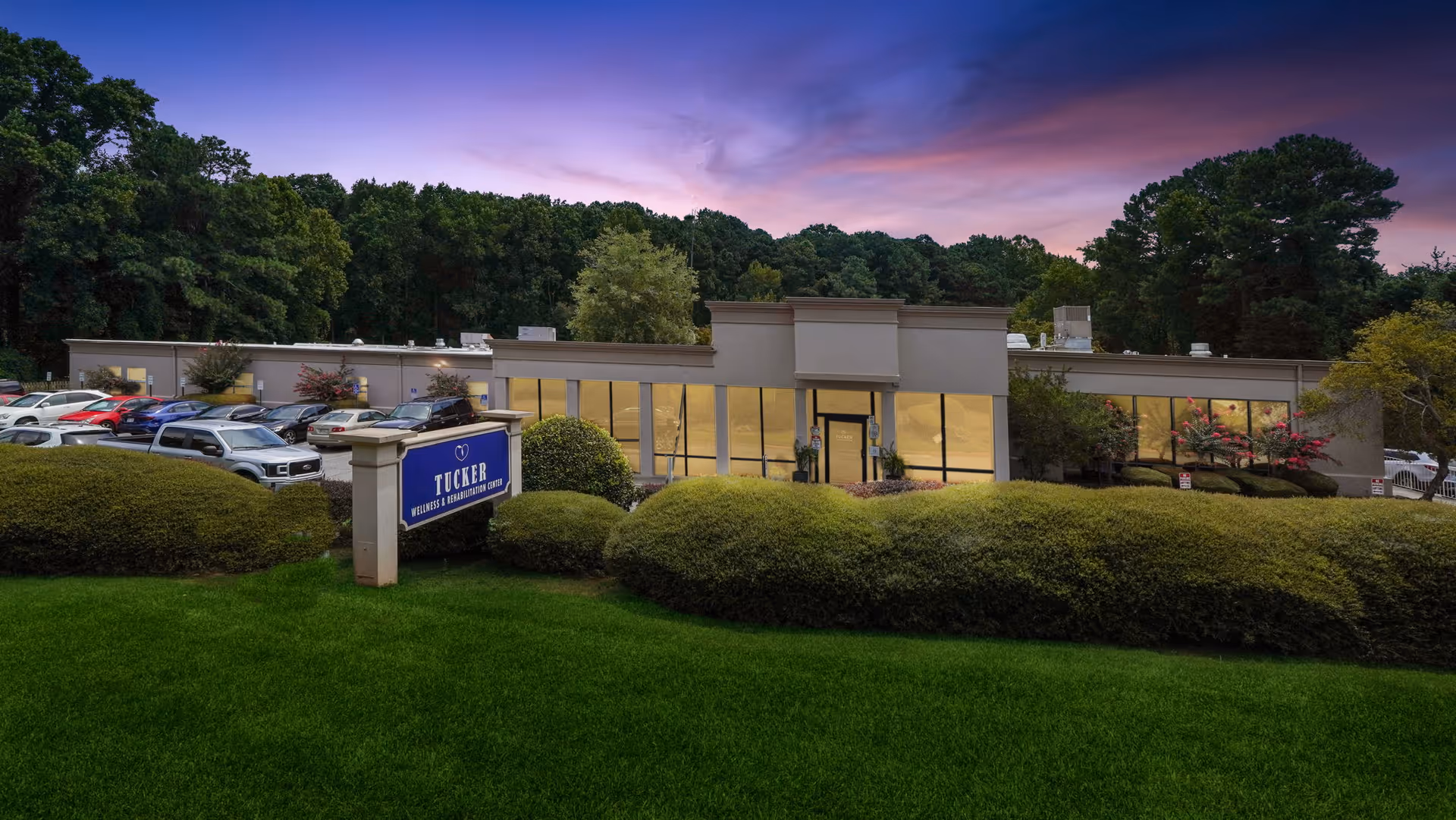 Exterior view of Tucker Nursing & Rehab Center building at dusk with a well-maintained lawn, bushes, and a parking lot with several cars. The sky is purple and pink, indicating sunset.