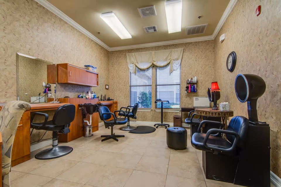 Interior of a salon room with multiple black salon chairs, a hair washing station, a hair dryer chair, a small table with a red lamp, and a large window with blinds and a valance. The walls have a textured beige wallpaper and the floor is tiled.