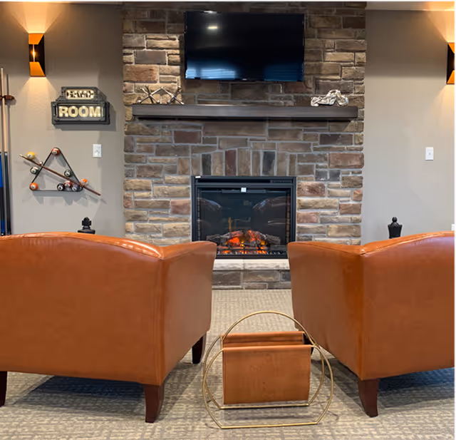 A cozy living room area with two brown leather armchairs facing a stone fireplace with a mounted flat-screen TV above it. The fireplace has a glowing fire, and there is a decorative magazine holder with a leather cover between the chairs. On the left wall, there is a wall-mounted pool ball rack and a sign that reads 'GAME ROOM'. Warm wall sconces provide lighting on both sides.