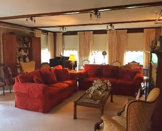 A cozy living room with two red sofas facing each other, a wooden coffee table with a floral centerpiece in between, beige carpet, several armchairs, and large windows with cream-colored curtains letting in natural light.