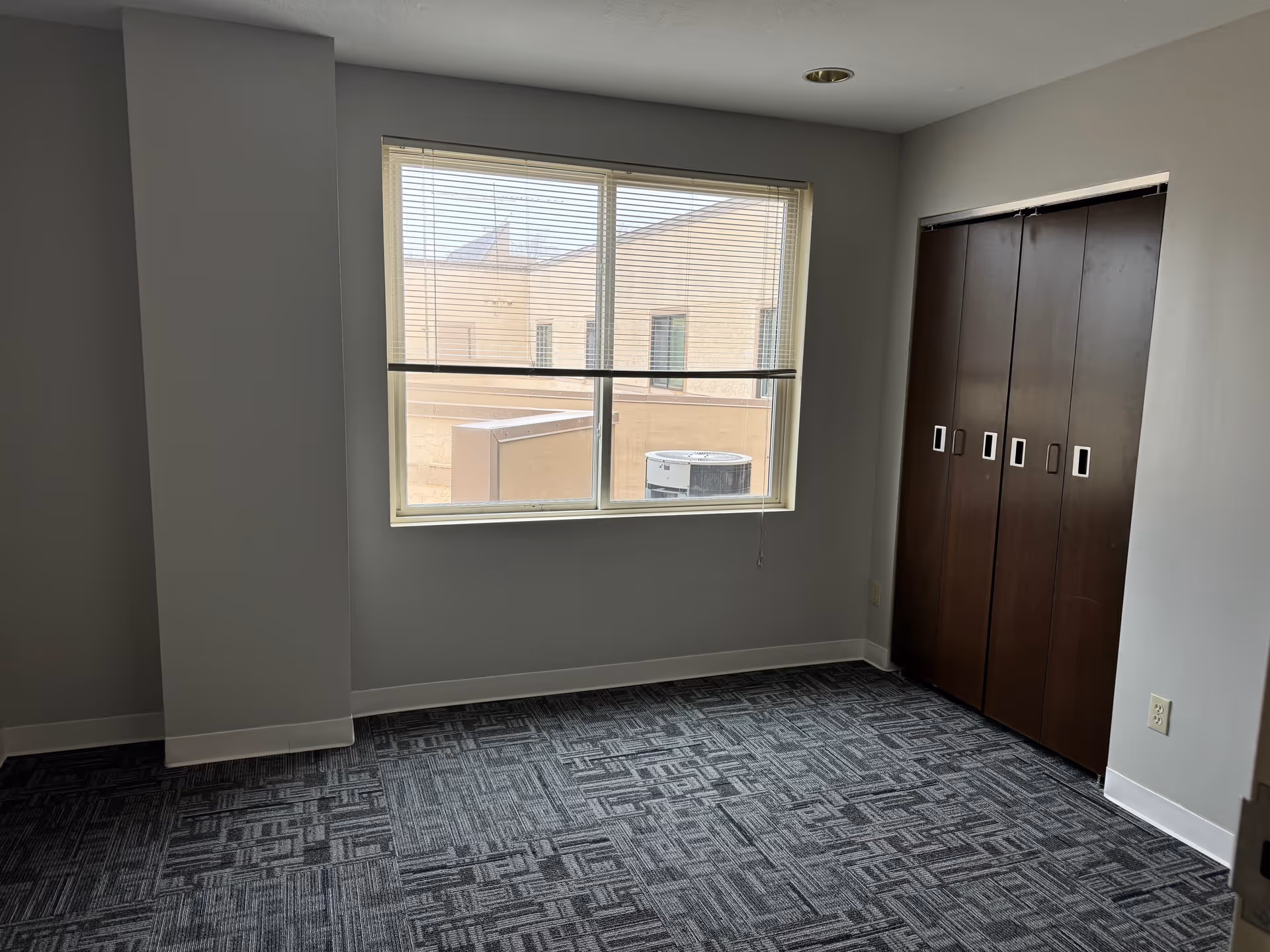 Empty room with gray patterned carpet, a large window with blinds partially open showing an exterior view of a building and HVAC unit, light gray walls, and a dark brown closet with four sliding doors.