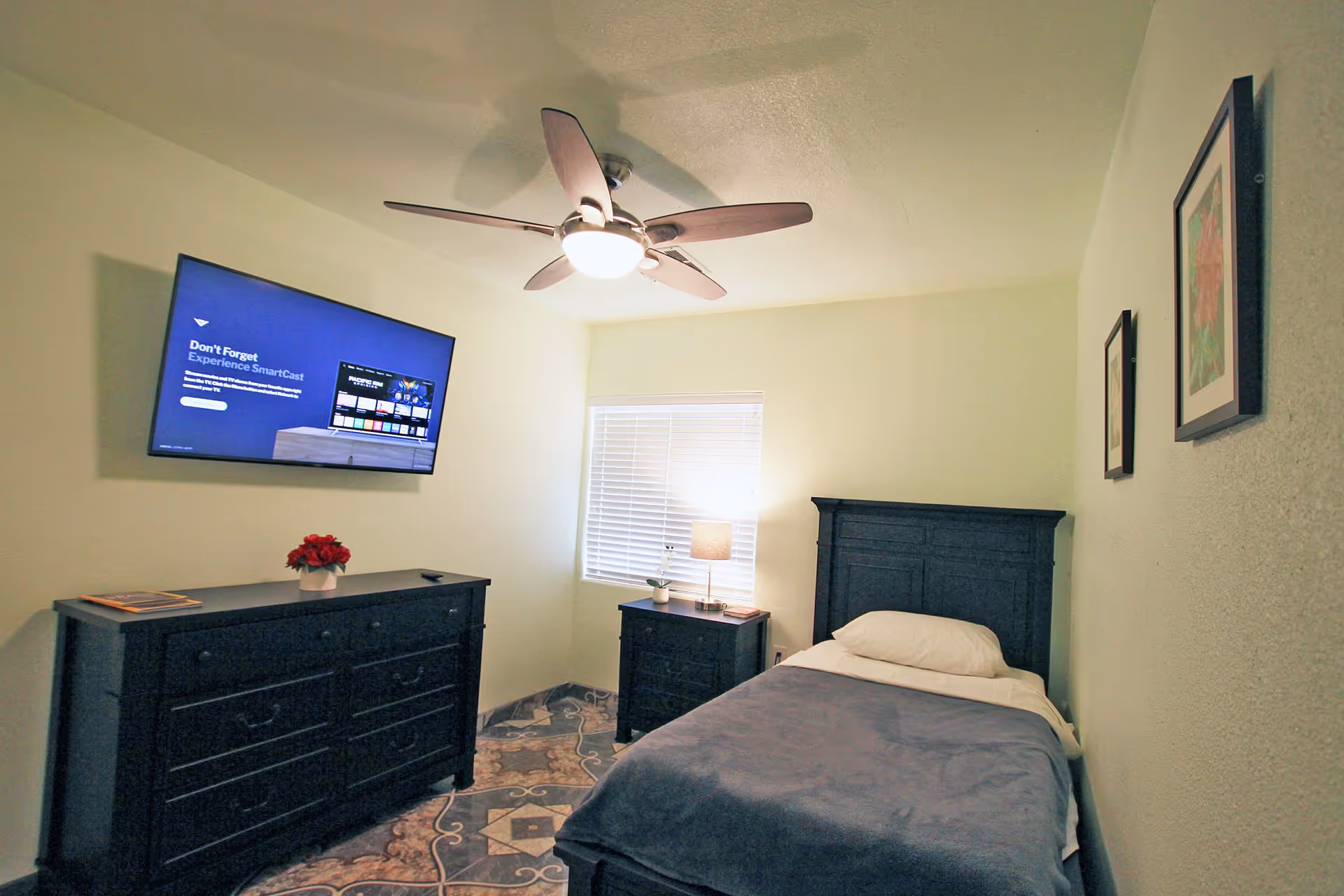 A small bedroom with a single bed covered with a blue blanket and a white pillow. There is a black wooden headboard, a matching black nightstand with a lamp and a small plant, and a black dresser with a red flower pot and a book on top. A flat-screen TV is mounted on the wall above the dresser. The room has a ceiling fan with a light, two framed pictures on the wall, and a window with closed blinds.