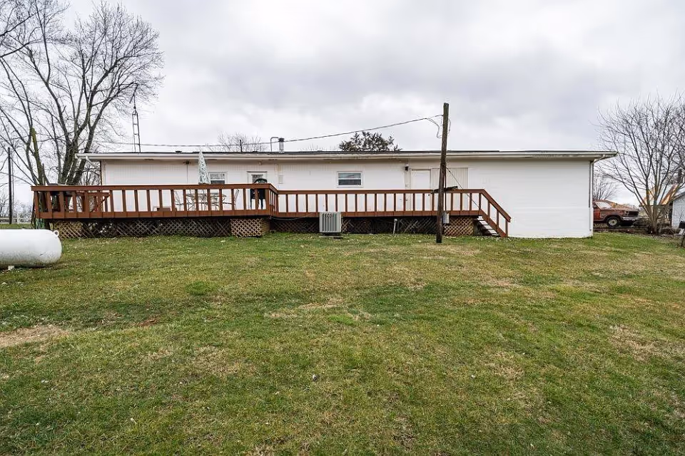 Exterior view of a single-story white building with a wooden ramp and stairs leading to the entrance. The building is surrounded by a grassy lawn with some leafless trees and a cloudy sky overhead.