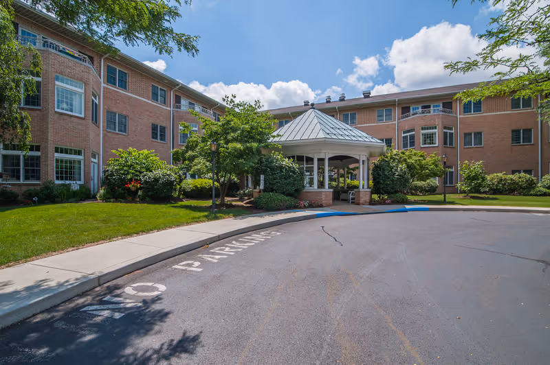 Exterior view of a senior living community building with a brick facade, multiple windows, and a covered entrance surrounded by greenery and trees under a blue sky with some clouds.