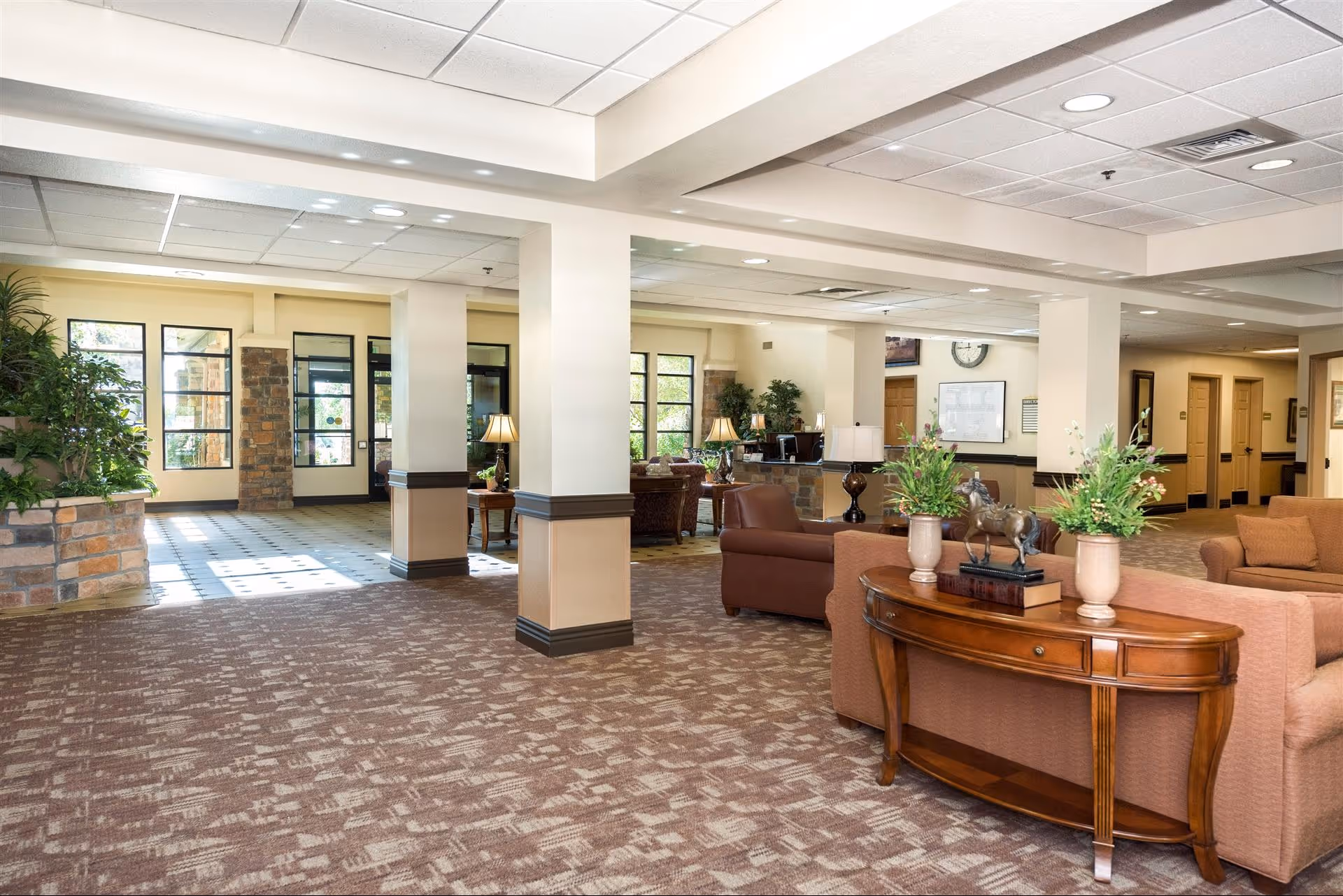 Spacious and well-lit senior living facility common area with beige walls, patterned carpet, and multiple seating arrangements including sofas and armchairs. There are decorative plants, table lamps, and a wooden console table with vases and a horse figurine. Large windows and glass doors allow natural light to enter the room.
