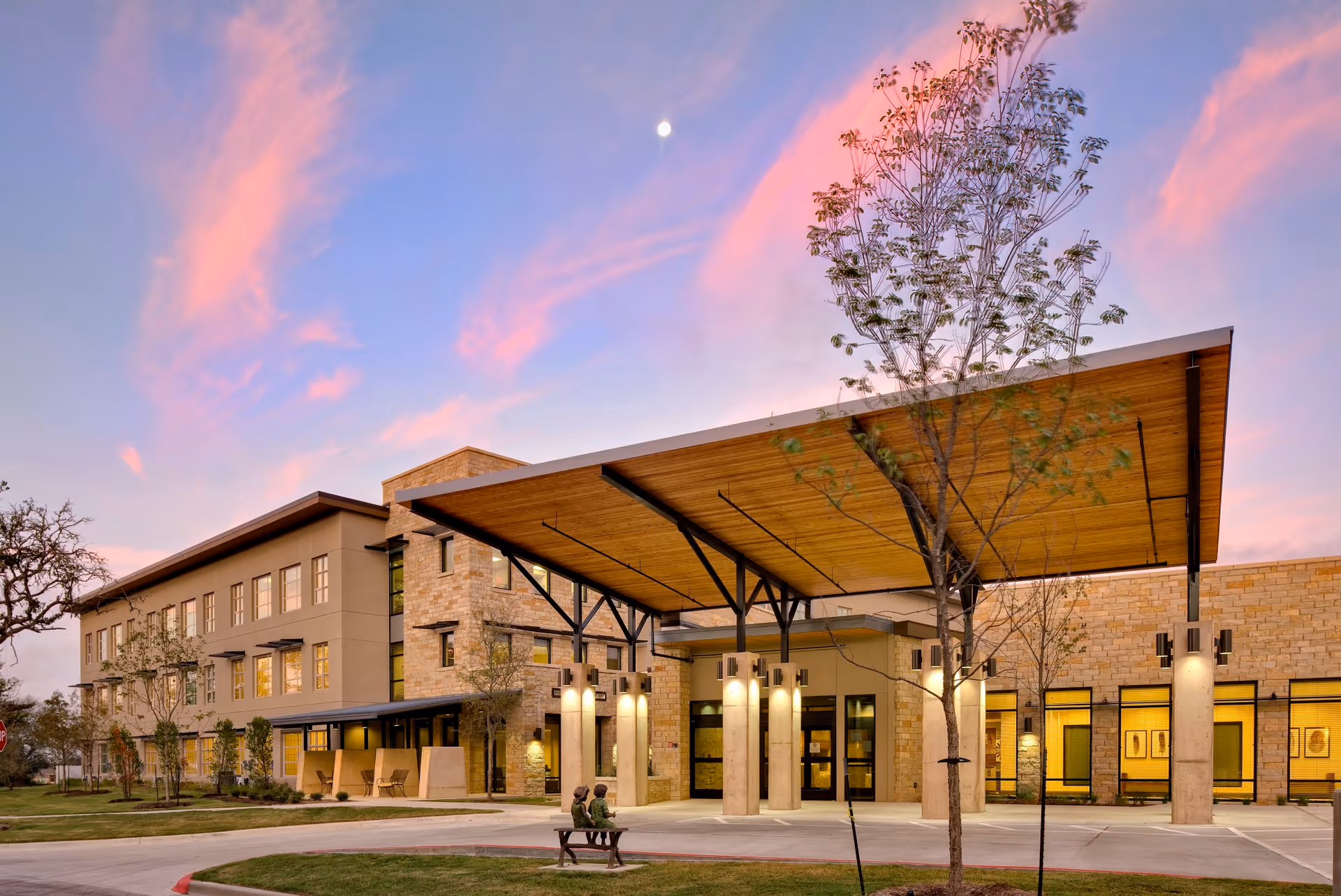 Exterior view of The Watermark at Southpark Meadows building at sunset with a large covered entrance supported by columns, trees, and a bench with two statues of children sitting on it in the foreground.