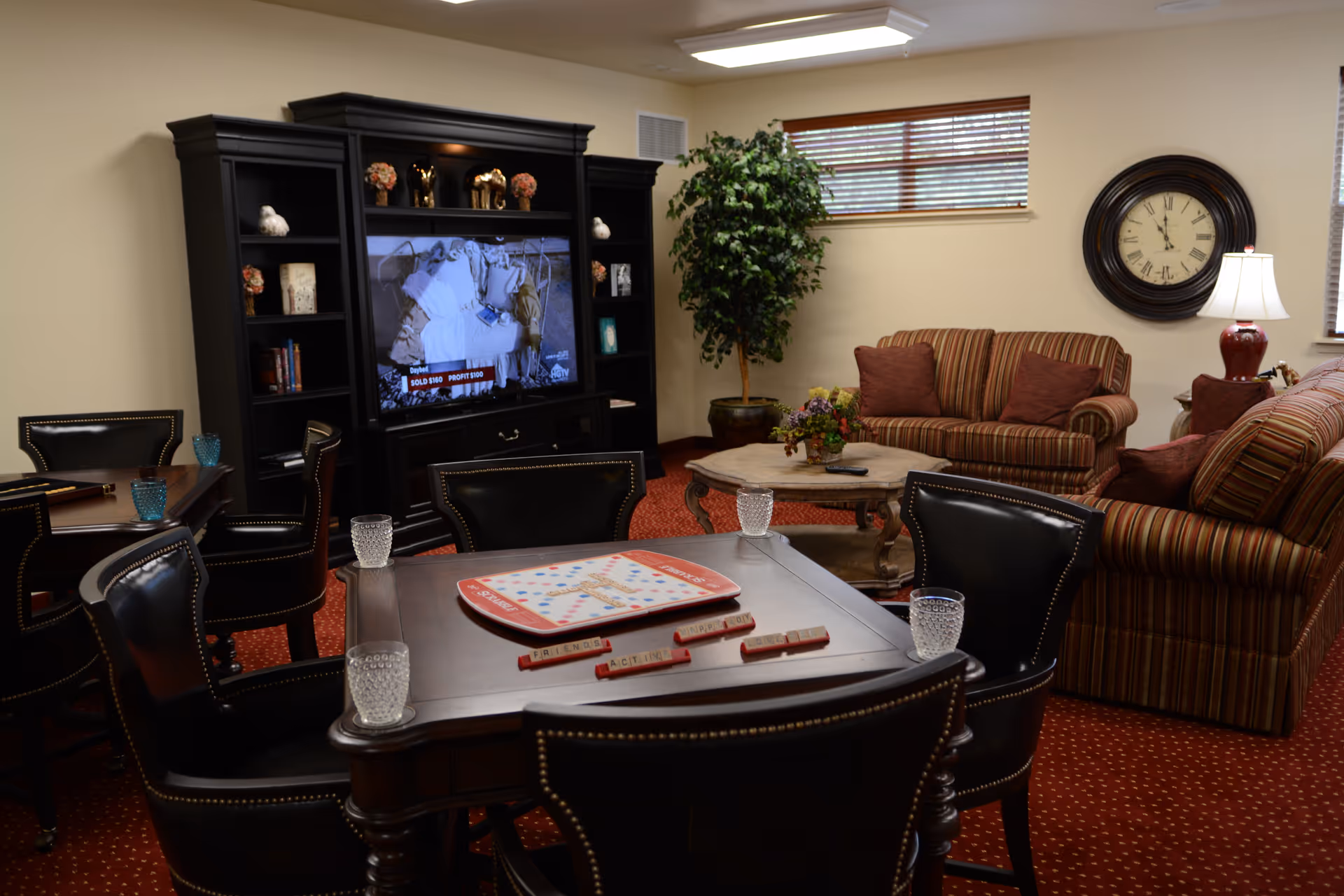 A cozy living room area with a dark wood entertainment center holding a television, two striped sofas with red and brown cushions, a round coffee table with a flower arrangement, a large wall clock, a table lamp, and a potted plant. In the foreground, there is a wooden game table with a Scrabble board and four black leather chairs around it, each with a glass of water on the table. The room has beige walls, two small windows with blinds, and a red carpet with a subtle pattern.