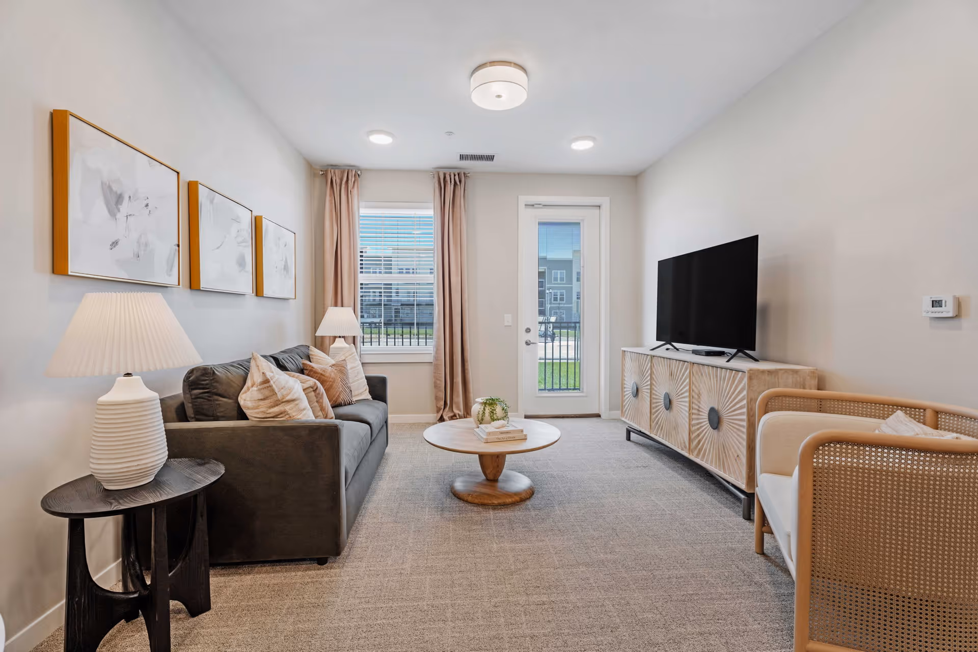 Bright neutral-toned living room with a gray sofa, round coffee table, TV on a console, and a glass door leading outside.