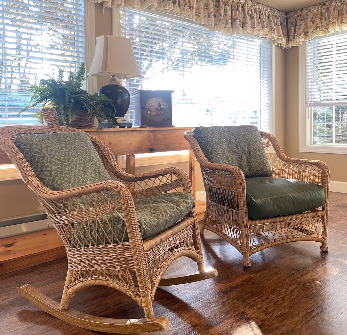 Two wicker chairs with green patterned cushions placed on a wooden floor in front of large windows with white blinds. Behind the chairs is a wooden console table with a black lamp, a potted plant, and a decorative box. The windows have floral valances at the top.