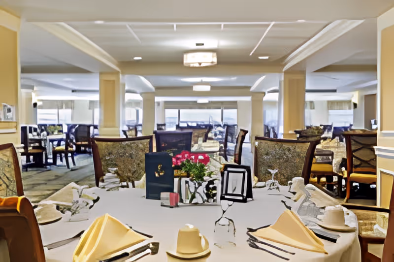A spacious dining room with multiple tables and chairs arranged neatly. The tables are set with white tablecloths, folded napkins, cups, and utensils. There are decorative centerpieces with flowers on the tables. Large windows in the background allow natural light to fill the room.