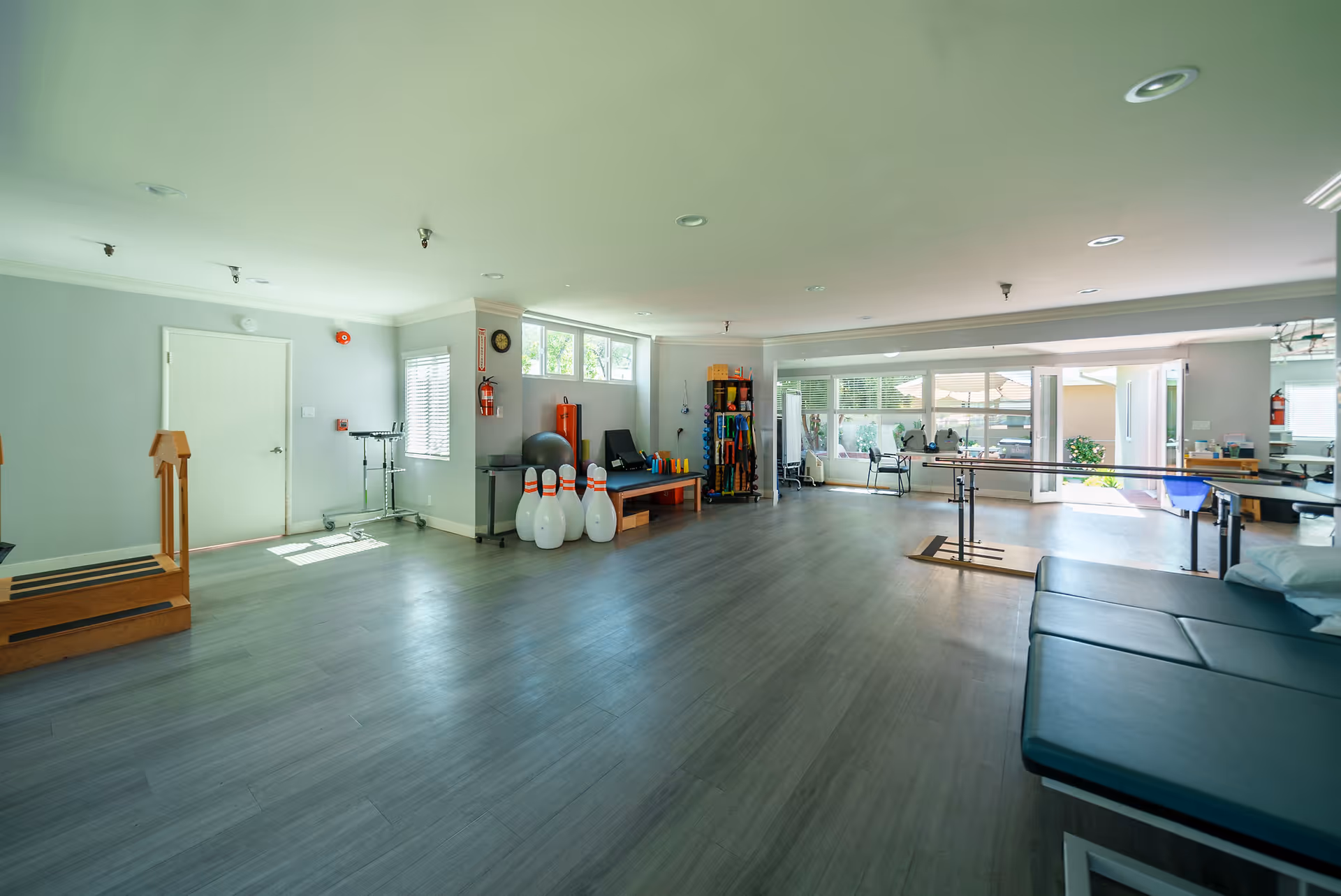 A spacious rehabilitation room with wooden stairs for physical therapy, exercise equipment including therapy balls, weights, and bowling pins, parallel bars for walking exercises, and large windows allowing natural light to fill the room.
