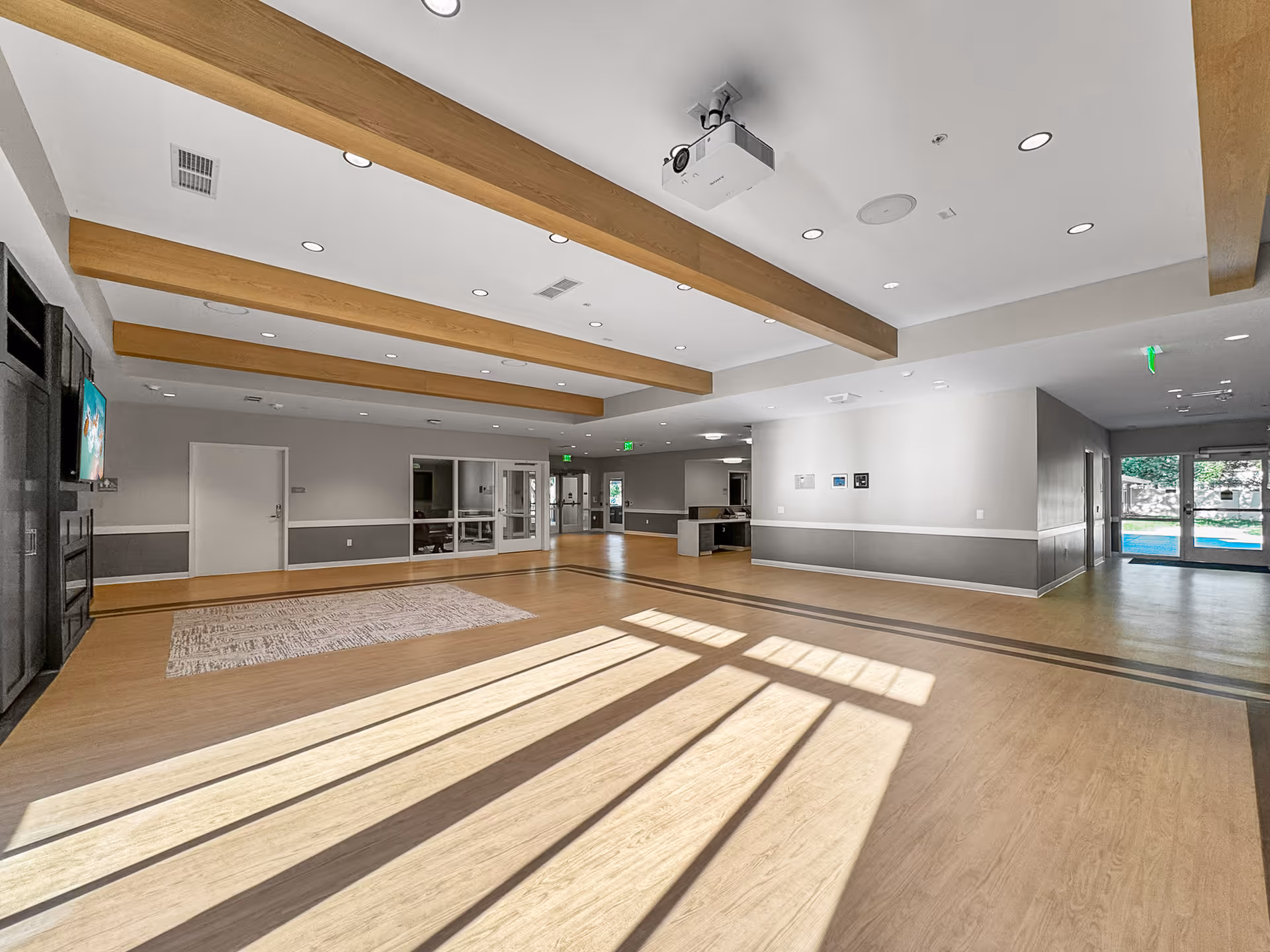 Spacious, bright multipurpose common room with exposed wood ceiling beams, large windows casting shadows on the floor, a wall-mounted TV, and doors leading outside.