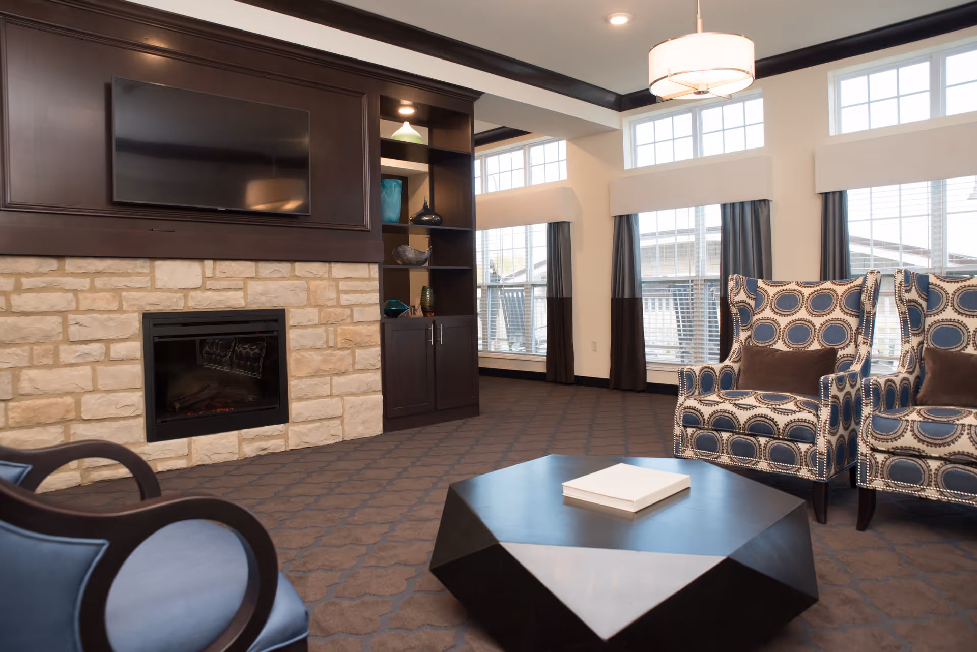 A cozy senior living apartment common area featuring a stone fireplace with a mounted flat-screen TV above it, dark wood built-in shelves with decorative items, large windows with white blinds and dark curtains, two patterned armchairs with brown cushions, and a modern black geometric coffee table with a book on top.