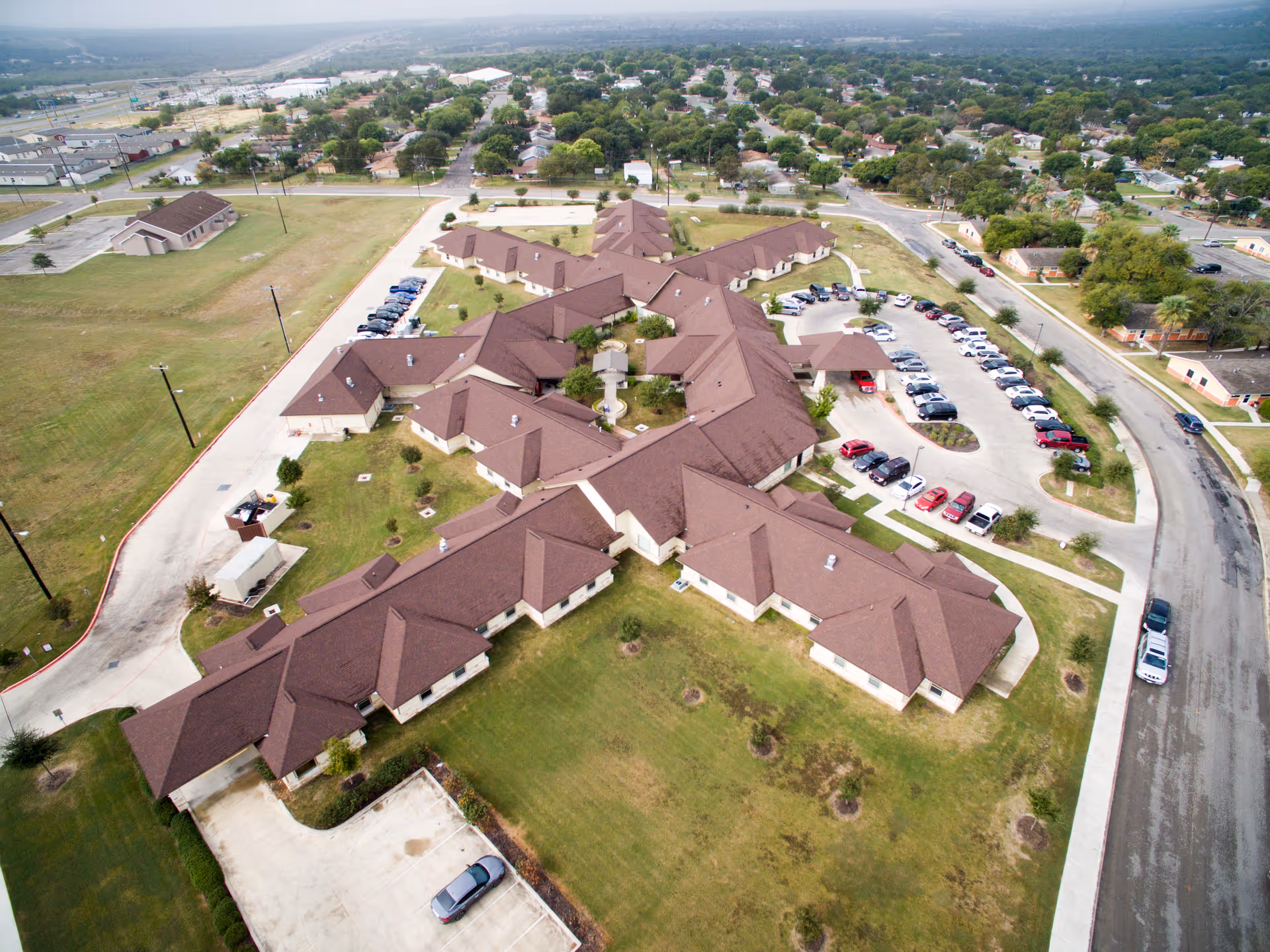 Aerial view of Legend Oaks Healthcare and Rehabilitation - West San Antonio, showing a large building complex with multiple wings and brown roofs surrounded by parking lots, green lawns, and nearby residential neighborhoods.