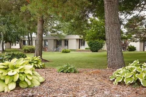 Exterior view of a single-story senior living facility with green lawn, trees, and landscaping.