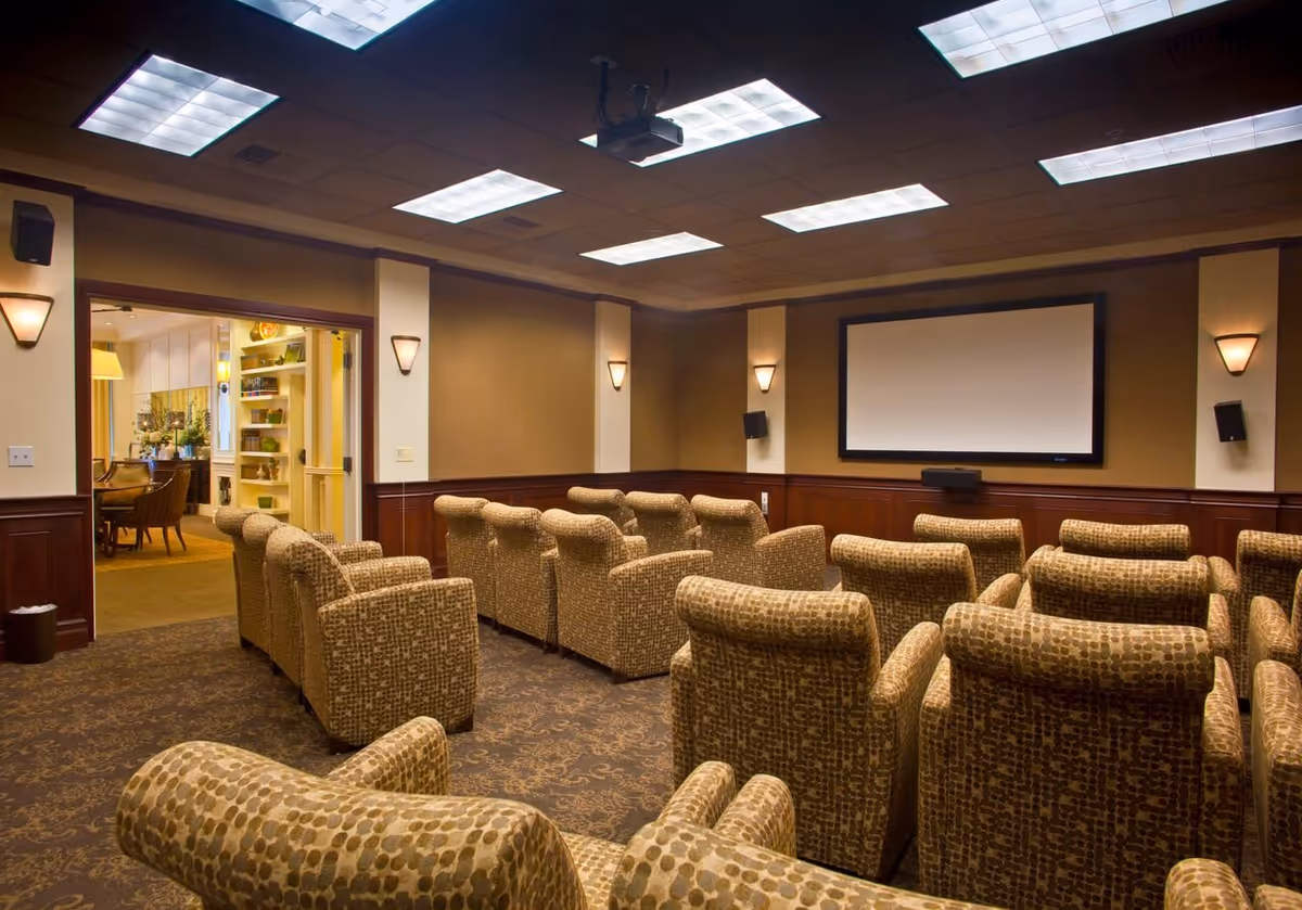 A cozy theater room with multiple rows of patterned upholstered armchairs facing a large blank projection screen on the wall. The room has warm lighting with wall sconces and ceiling lights, and an open doorway leads to an adjacent room with shelves and additional seating.