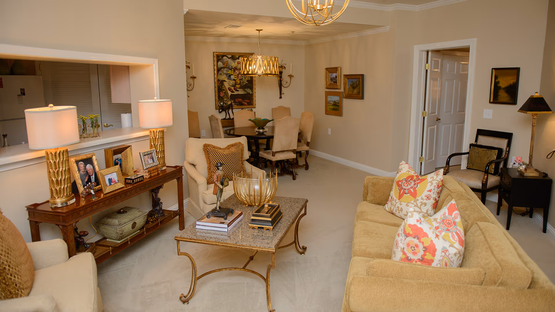 A cozy living room and dining area in a senior living facility. The living room features a beige sofa with floral patterned pillows, an armchair with a decorative pillow, a coffee table with books and decorative items, and a side table with framed photos and two lamps. The dining area has a round table with six upholstered chairs, a hanging light fixture, and framed artwork on the walls. The walls are painted a warm beige color, and the carpet is light-colored. There is a doorway leading to another room and a pass-through window to the kitchen.