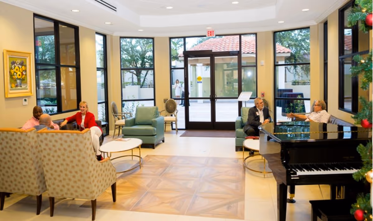 A bright and spacious senior living facility common area with large windows and glass doors letting in natural light. The room features comfortable seating including armchairs and sofas arranged around small coffee tables. Two groups of people are engaged in conversation. A black grand piano is visible on the right side near a decorated Christmas tree. Outside, trees and a tiled roof structure can be seen through the glass doors.