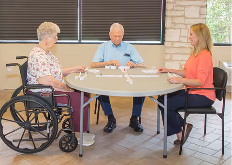 Three people sitting around a round table playing a game of dominoes. One elderly woman in a wheelchair, an elderly man, and a younger woman are engaged in the game in a well-lit room with a stone wall and window blinds in the background.