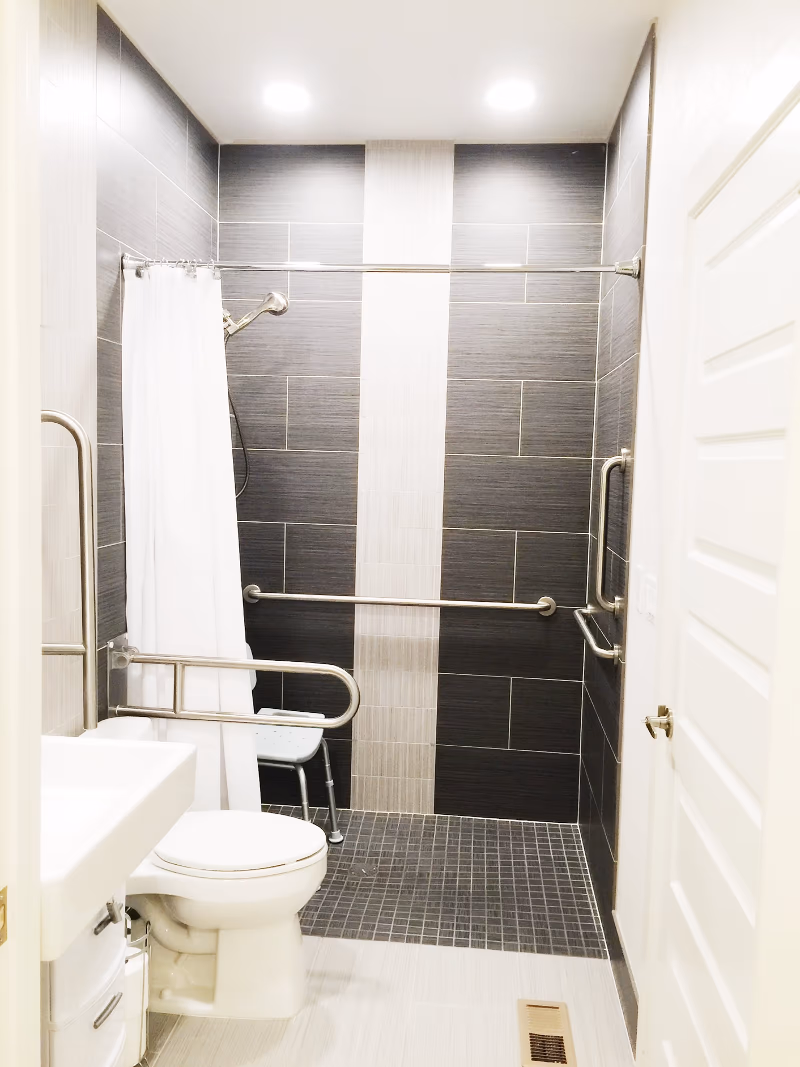 A modern accessible bathroom with dark gray and light beige tiled walls and floor. It features a white toilet, a white sink, a shower area with a white curtain, a shower chair, and multiple stainless steel grab bars for support.