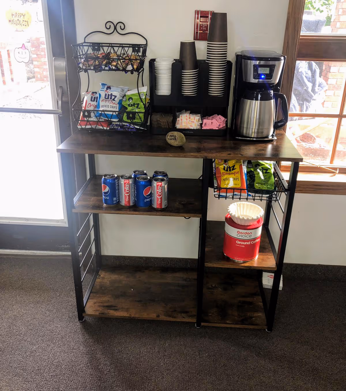 A wooden and metal shelving unit holding a coffee maker, disposable cups, sugar and sweetener packets, various bags of Utz potato chips, cans of Pepsi and Diet Coke, and a canister of Gordon Choice ground coffee with coffee filters on top. The unit is placed near a door and a window with a view outside.