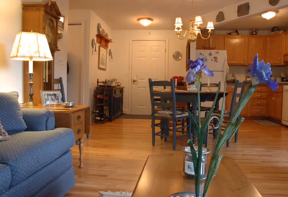 A cozy living and dining area with a blue upholstered armchair on the left, a wooden side table with a lamp and framed photo, a wooden dining table with chairs in the center, and a kitchen with wooden cabinets and a white refrigerator in the background. Purple flowers in a vase are on a wooden coffee table in the foreground.