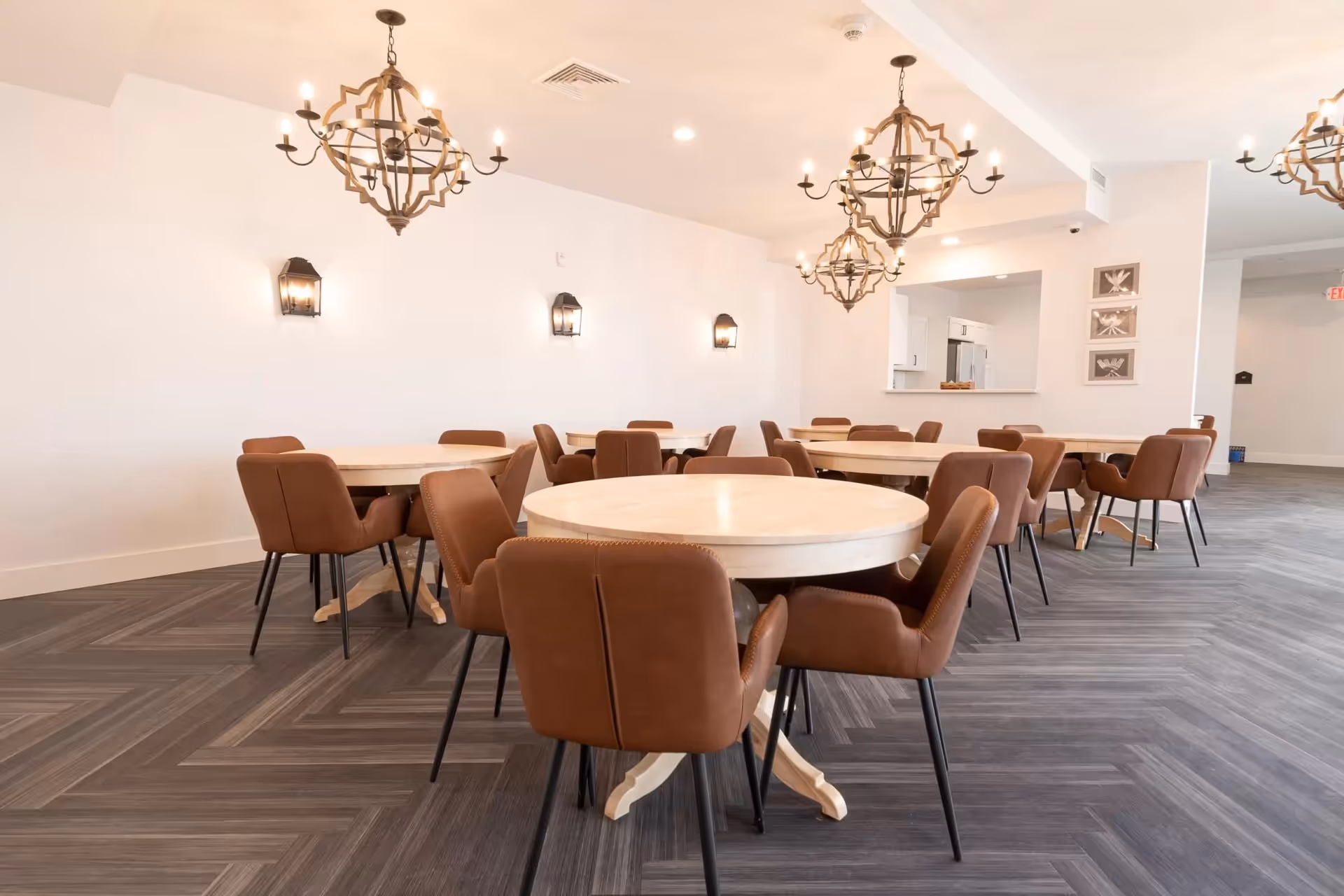 A bright dining room with multiple round wooden tables surrounded by brown cushioned chairs. The room features modern chandeliers hanging from the ceiling and wall-mounted lantern-style lights. The floor has a dark herringbone pattern, and there is a pass-through window to a kitchen area in the background.