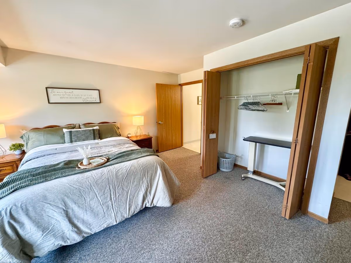 A cozy bedroom with a neatly made bed featuring green and gray bedding. There are two wooden nightstands with lamps on either side of the bed. Above the bed is a framed sign with a quote. To the right, there is an open closet with wooden bi-fold doors, containing hangers, a laundry basket, and an ironing board. The room has beige walls and carpeted flooring.